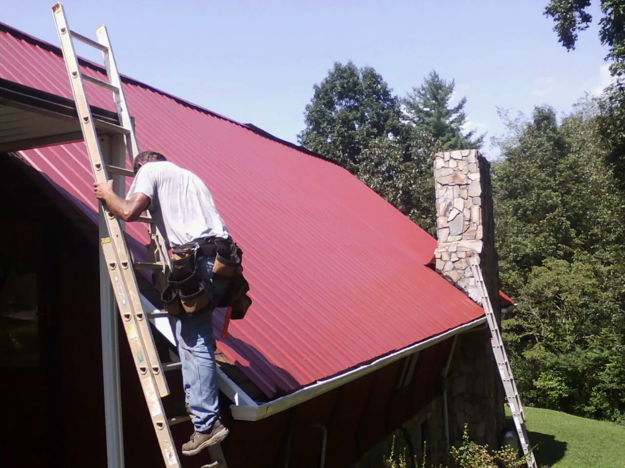Roofer on a ladder beside a red metal roof and stone chimney — Soffit & Fascia Repair by Roofing Brothers in Paxinos