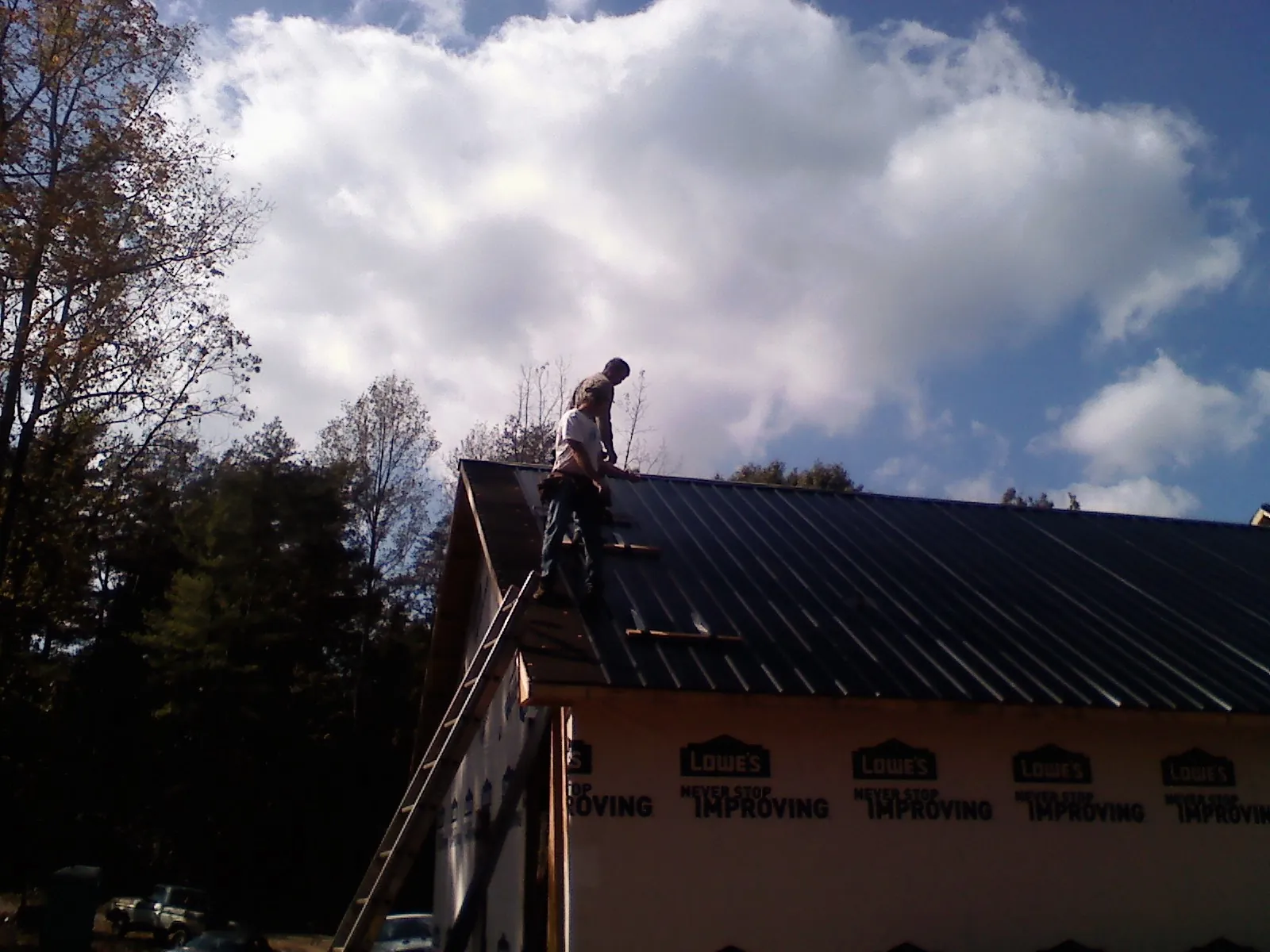 Roofer fastening dark metal panels high against a cloudy sky, from a Roof Inspection job in Paxinos